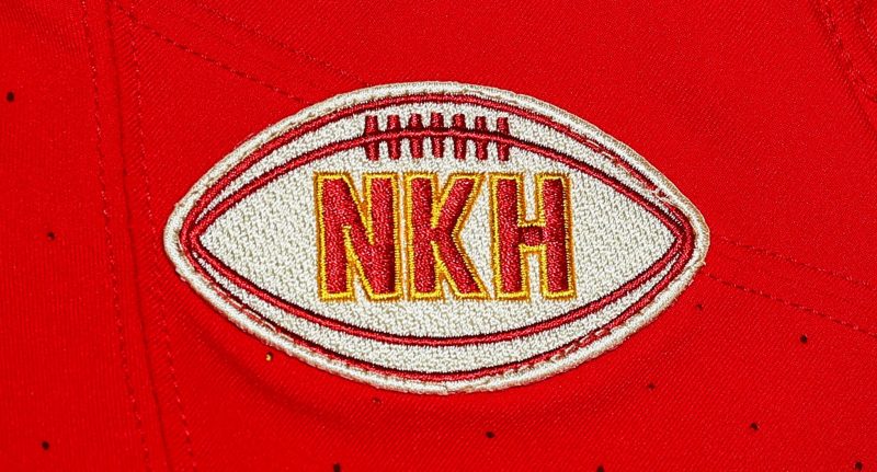 Patch dei Kansas City CHiefs in ricordo di Norma Knobel Hunt, moglie del fondatore Lamar Hunt. Le iniziali NKH sono in rosso bordate di giallo e sono posizionate all'interno di una palla da football